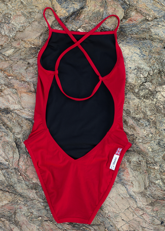 A red one-piece swimsuit with a black center panel and crisscross back straps, laid flat on a textured gray and brown stone surface. A small white tag with a red Union Jack and the brand name 'MACKE' is visible near the lower right side.